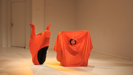 Zwei Performerinnen befinden sich jeweils in einem orangenen Stofflaken. Die Performerin auf der linken Seite ist gar nicht zu sehen. Dennoch kann man ihren Umriss unter dem Laken erkennen, da sie eine Pose einnimmt, durch die das Laken gespannt ist.