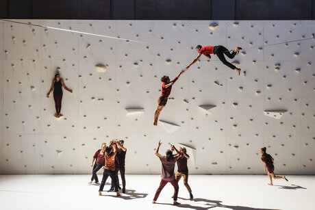 Performer:innen befinden sich an und auf einer Kletterwand, darüber liegt ein Performer auf einem quer über die Bühne gespannten Seil.