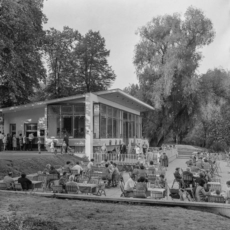 Schwarz-Weiß-Fotografie des Milchhäuschens in Berlin-Weißensee, vor dem viele Menschen in kleinen Grüppchen an Tischen im Außenbereich sitzen. Das Milchhäuschen wird von hohen Bäumen gesäumt.