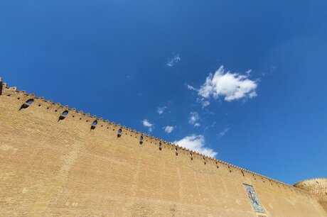 Das Bild zeigt eine hohe mit schmuckvollen Zinnen versetzte Mauer, die gegen einen strahlend blauen Himmel mit wenigen Schönwetterwolken fotografiert ist.