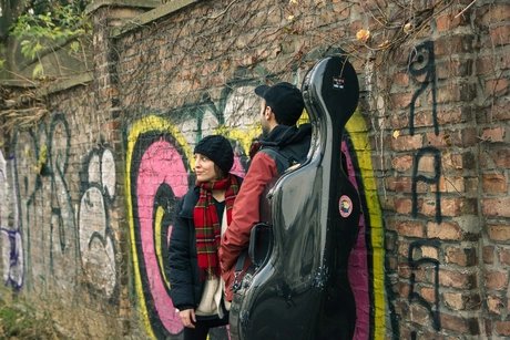 Zwei Personen stehen vor einer Backsteinwand, die mit Graffiti besprayt ist. Bei der linken Person handelt es sich um die Gastgeberin Antonia Regis. Die rechte Person ist nur von hinten zu sehen, sie trägt ein Cello-Koffer.