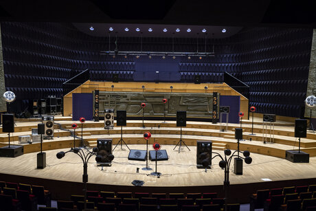 Mehrere Lautsprecher in unterschiedlichen Formen und Größen stehen auf einer großen Bühne. Der Publikumssaal ist leer.