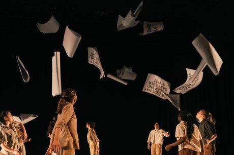 Eine Gruppe junger Menschen, in Beige- und Brauntönen gekleidet, steht auf einer Bühne. Durch die Luft fliegen Zettel mit schwarzer Beschriftung.