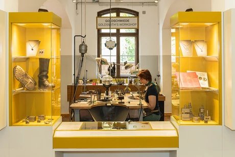 Zwischen zwei gelben Vitrinen mit unterschiedlichen Objekten blickt man auf einen Tisch mit zahlreichen, verschiedenen Goldschmiede-Geräten. An dem Tisch sitzt eine Frau und arbeitet.