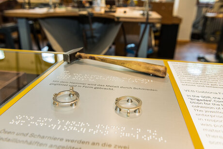 Auf einem Ausstellungstisch sind ein Hammer sowie zwei Ringe zum Tasten befestigt. Darunter ist Braille Schrift angebracht.