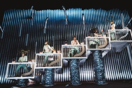 Fünf Personen performen auf fünf ansteigenden Plateaus aus Plexiglas; um sie herum glatte Röhren und geriffelte Rohre.