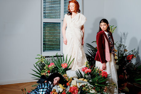 Drei rätselhafte Frauen um ein Blumenarrangement herum, blicken in verschiedene Richtungen