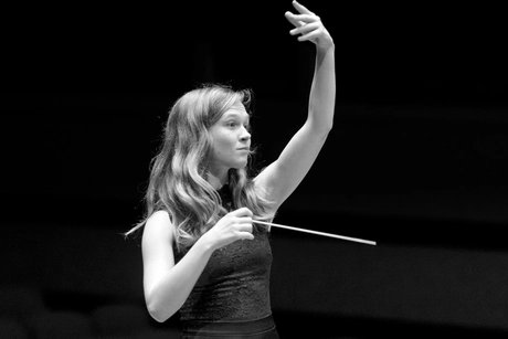 Dirigentin Mirga Gražinytė-Tyla hält in der rechten Hand den Taktstock und hebt den linken Arm vor dem Orchester hoch.