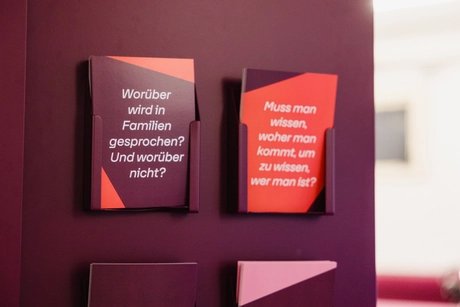 Zwei Informationskarten in Haltern an einer dunkelroten Wand, eine dunkelrot mit weißem Text 'Worüber wird in Familien gesprochen? Und worüber nicht?', die andere rot mit weißem Text 'Muss man wissen, woher man kommt, um zu wissen, wer man ist?'.