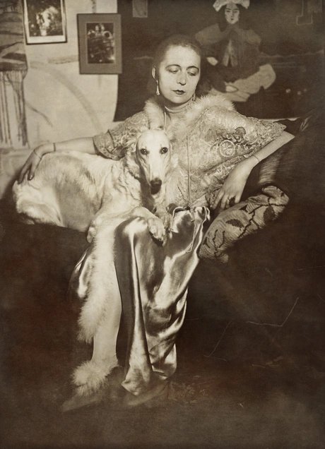 Selbstportrait der Künstlerin Marta-Astfalck-Vietz als Sepia-Fotografie. Sie sitzt in einem glänzenden Kleid mit Pelzbesatz auf einem Sofa. Auf ihrem Schoß sitzt ein Hund mit hellem Fell. Im Hintergrund sind Gemälde und dekorative Elemente zu sehen.