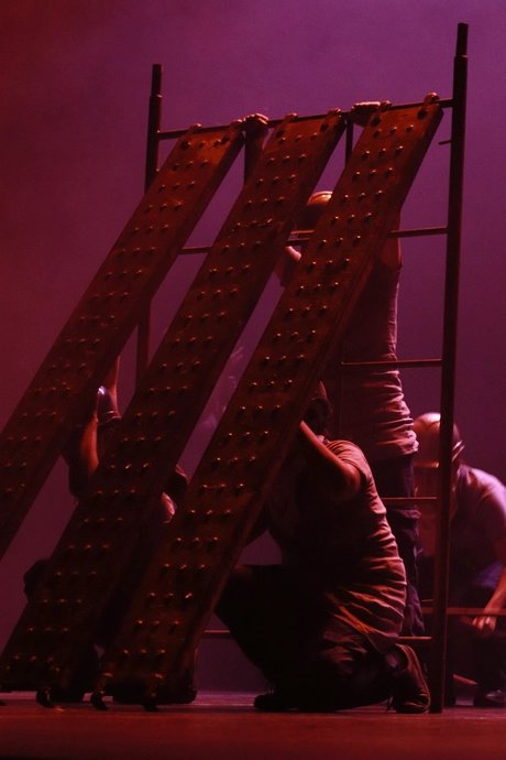 Mehrere Performer*innen halten große, rechteckige Metallplatten in einer schrägen Konstruktion. Die Szene ist in rotes und violettes Bühnenlicht getaucht. Körper und Gesichter sind teilweise verdeckt.