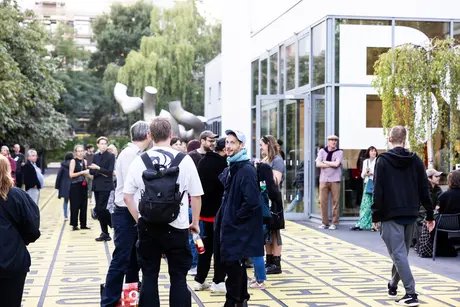 Auf dem Vorplatz der Berlinischen Galerie stehen einige Menschen in kleineren Gruppen zusammen und unterhalten sich miteinander. Eine Person blickt direkt in die Kamera.