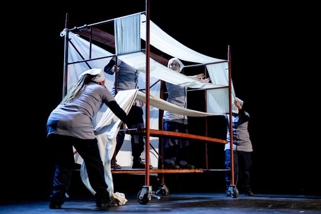 Mehrere Performer*innen bewegen sich um ein rollbares Gerüst auf der Bühne. Weiße Stoffbahnen sind über die Konstruktion gespannt und werden von den Beteiligten gehalten und arrangiert. Die Bühne ist dunkel, das Licht konzentriert sich auf die Szene.