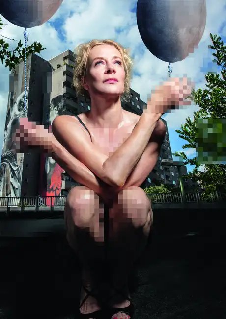 Katja Riemann sitzt unbekleidet in der Hocke vor einem Plattenbau und hält jeweils einen gräulichen Luftballon in den Händen. Die Hände, der Brust- und Intimbereich sind verpixelt.