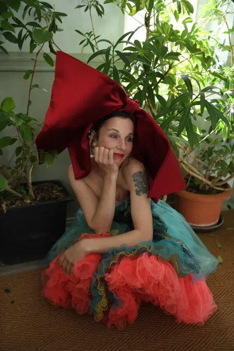 Meret Becker sitzt in einem Kleid aus türkis- und rosa-farbenem Stoff mit einer großen roten Schleife auf dem Kopf in der Hocke. Der Hintergrund zeigt verschiedene Zimmerpflanzen.