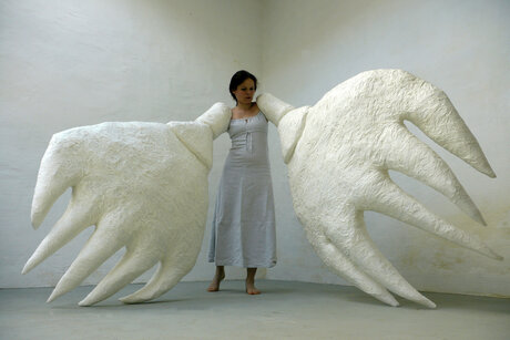 Ilka Raupach steht im Zentrum eines leeren Raumes. An beiden Armen trägt sie aus Papier gefertigte Hände in übermenschlicher Größe.