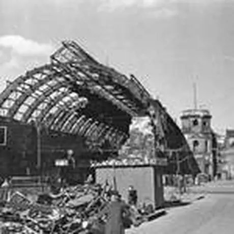 Blick entlang der Straße, links die zersplitterte Bahnhofshalle und der ausgebrannte Bahnhofsturm, rechts die fensterlosen Fassaden beschädigter Wohnhäuser