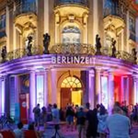 Das Museum bei Nacht von außen mit bunter Leuchtschrift, auf der BERLINZEIT steht, davor Menschen