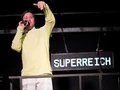 Eine Person in einem gelben Pulli und mit einer weissen Hose redet in ein Mikrophon. Hinter ihm hängt ein Schild auf dem Superreich steht.