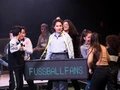 Eine Person steht in der Mitte des Bildes und um ihren Hals hängt ein Schild auf dem Fußballfans steht. Um die Person herum stehen mehrere Kinder.