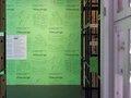 Raumbild mit auf beiden Seiten Schränken mit Vitrinen und Regale mit Umzugskartons. Im Hintergrund befindet sich eine mit grell grünen Plakaten tapezierte Wand mit den Keyvisuals: Gebiss, Hund-Luftballon, Karo-Tasche und Kurzhanteln.