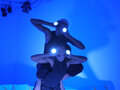 Der Raum ist in blaues Licht getaucht. Zwei Performerinnen halten sich die Hände vor die Augen. Auf ihren Handrücken befinden sich runde, leuchtende Kreise. Die eine Performerin sitzt aufrecht auf dem Rücken der anderen.