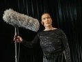 Frau in glitzerndem schwarzen Outfit hält ein großes, mit Fell verkleidetes Mikrofon und schaut konzentriert nach vorn.