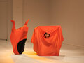 Zwei Performerinnen befinden sich jeweils in einem orangenen Stofflaken. Die Performerin auf der linken Seite ist gar nicht zu sehen. Dennoch kann man ihren Umriss unter dem Laken erkennen, da sie eine Pose einnimmt, durch die das Laken gespannt ist.
