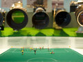 Blick in eine Vitrine: Im Vordergrund liegt ein Fußballfeld aus grünem Filz, auf dem Miniaturfiguren einem Tor entgegen stürmen. Im Hintergrund sind fünf verschiedene Foto-Objektive auf die Szene gerichtet.