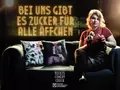 Postkartenmotiv zur Veranstaltung. Im oberen Bereich steht in goldleuchtenden Neonbuchstaben: "Bei uns gibt es Zucker für alle Äffchen". Darunter ein Sofa, auf dem die Gastgeberin Nicki mit einem Glas Rotwein in der Hand sitzt. In der anderen Hand hä