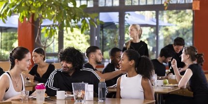 Un grupo de personas está sentado en las mesas de una cafetería muy iluminada del hotel Citylight. Charlan y disfrutan de la comida. Una mujer observa desde el fondo. Los grandes ventanales proporcionan luz natural.