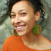 Portrait-Foto der lächelnden Jessica Cuna in organgefarbenem Pullover und mit großen, runden, grün-orangen Ohrhängern