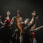 Acht Performer:innen stehen eng beisammen, die Arme anmutig erhoben, rote und grüne Bänder in den Händen – ein kraftvoller, ritueller Moment in dramatischem Bühnenlicht.