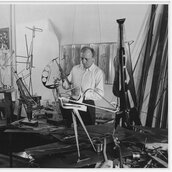Schwarz-Weiss-Fotografie von Hans Uhlmann in seinem Atelier. Um den Künstler herum sind viele Metall-Skulpturen im Entstehungsprozess zu sehen.