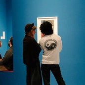 Rückenansicht von zwei Personen, die vor einer Collage von Raoul Hausmann stehen, die an einer leuchtend blauen Wand hängt.