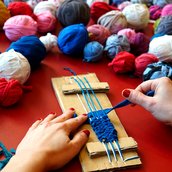 Eine Person sitzt an einem Tisch und webt an einem selbstgebauten Webrahmen aus Pappe aus einem breiten, blauen Stück Stoff ein Band. Von der Person sind nur die Hände zu sehen. Auf dem Tisch liegen viele bunte Knäule aus Stoffresten.