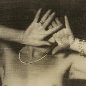Körnige Schwarz-Weiß-Fotografie: Porträt einer unbekleideten Person mit Perlenkette, die beide Hände vor das Gesicht hält, sodass dieses nicht sichtbar ist. Die Hände tragen Schmuck, darunter ein Ring und ein Armband.