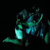 Zwei Performer:innen in intensiver Pose auf dunkler Bühne, grünlich beleuchtet, mit ineinander verschlungenen Körpern.