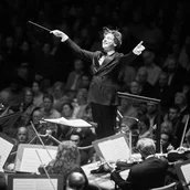 Dirigent Klaus Mäkelä breitet die Arme vor dem Orchester aus, die Musiker:innen streichen ihre Geigen und im Hintergrund sieht man das Publikum.