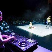 Menschen tanzen auf einer beleuchteten Bühne in einem sonst dunklen im Raum. Im Vordergrund ist angeschnitten ein*e DJ an Turntables zu sehen.
