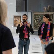 Aya und Michel tragen rot-blaue Schals des Jüdischen Museums Berlin und stehen dort in der Dauerausstellung. Sie unterhalten sich in Deutscher Gebärdensprache mit den Besuchern.