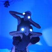 Der Raum ist in blaues Licht getaucht. Zwei Performerinnen halten sich die Hände vor die Augen. Auf ihren Handrücken befinden sich runde, leuchtende Kreise. Die eine Performerin sitzt aufrecht auf dem Rücken der anderen.