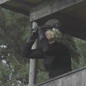 Ein blonde Frau beobachtet auf einem Hochsitz im Wald etwas mit einem Fernglas.