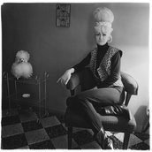 Eine Person sitzt auf einem Stuhl und hat die Beine übereinander geschlagen. Neben ihr steht ein Regal mit einem künstlichen Souvenirhund darauf.