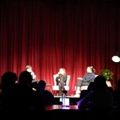 Hinter den Schatten des Publikums sieht man verschwommen drei Personen auf der Bühne vor einem roten Vorhang. Sie sitzen auf Stühlen und halten Mikrofone.