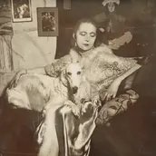 Selbstportrait der Künstlerin Marta-Astfalck-Vietz als Schwarz-Weiß-Fotografie. Sie sitzt in einem glänzenden Kleid mit Pelzbesatz auf einem Sofa. Auf ihrem Schoß sitzt ein Hund mit hellem Fell. Im Hintergrund sind Gemälde und dekorative Elemente zu 
