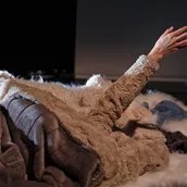 Auf dem Bild zu sehen ist ein kleiner Haufen aus verschiedenen Pelzen und einem Schaffell auf einer Bühne. Daraus ragt, offenbar auch in einen dieser Pelze gekleidet, der Arm einer Person, deren Körper wohl unter den Pelzen liegt. Die Pelze sind, bis