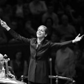Dirigentin Karina Canellakis breitet die Arme vor dem Orchester aus, die Musiker*innen streichen ihre Celli und im Hintergrund sieht man das Publikum.