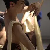 Mehrere Performer:innen mit nacktem Oberkörper und traditionell anmutenden Stoffbahnen in sanften, fließenden Bewegungen; Fokus auf eine zentrale Person mit erhobenen Armen in meditativer Pose.