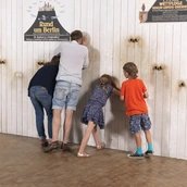 Eine vierköpfige Familie steht vor einer weißen Wand. Durch Gucklöcher in der Wand werfen sie einen Blick auf dahinter angebrachte Fotografien historischer Flugplätze. Auf der Wand sind historische Werbeplakate für Wettflüge aufgehängt.